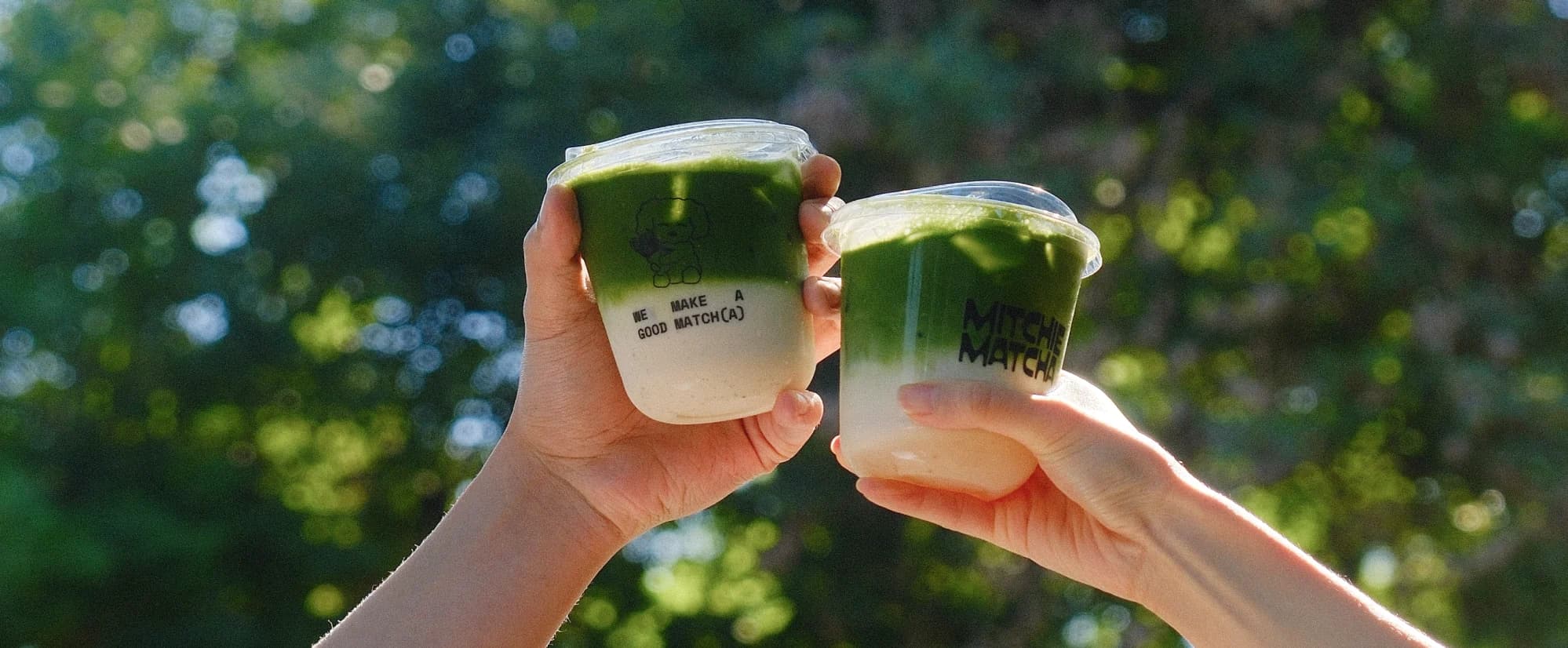 Two hands raising Mitchie Matcha iced matcha lattes in a toast against green trees