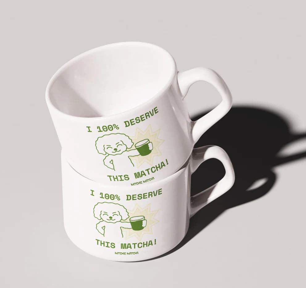 Two stacked white mugs printed with a green illustration and the words “I 100% deserve this matcha!”