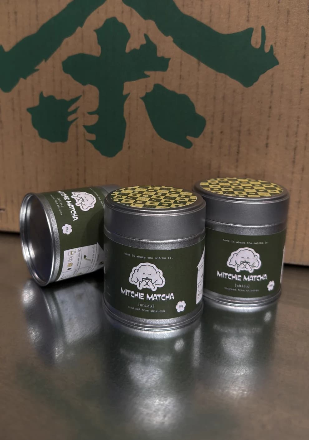 Three Mitchie Matcha tins on a counter in front of a green tea character sign
